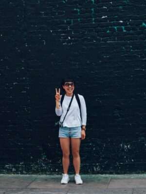 Smiling woman in sunglasses, posing with a peace sign against a brick wall.